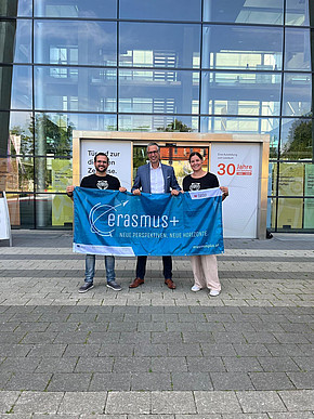 Stehen mit der Flagge der ErasmusDays2025 in der Hand vor der FH Salzburg: Kevin Bailey, FH Rektor Dominik Engel, Jennifer Simon