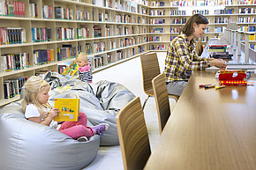 Zwei Kinder spielen im Vordergrund, Person arbeitet seitlich am Tisch